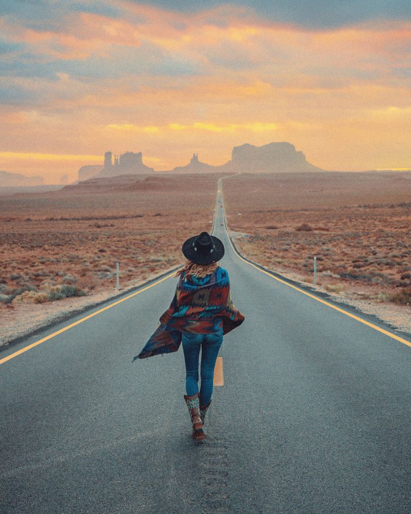 Une femme marchant sur une route désertique en direction de Monument Valley avec coucher de soleil spectaculaire